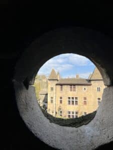 Vue d'un bâtiment médiéval à tours et fenêtres rouges à travers une meurtrière circulaire en pierre.
