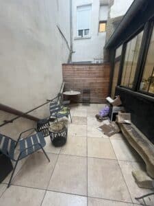 Petite terrasse pavée avec mobilier de jardin en métal, mur en bois et encombrants près d'une grande verrière.