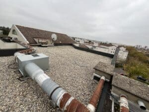 Toit terrasse gravillonné avec conduits de ventilation rouillés, antenne satellite et vue urbaine sous ciel couvert.